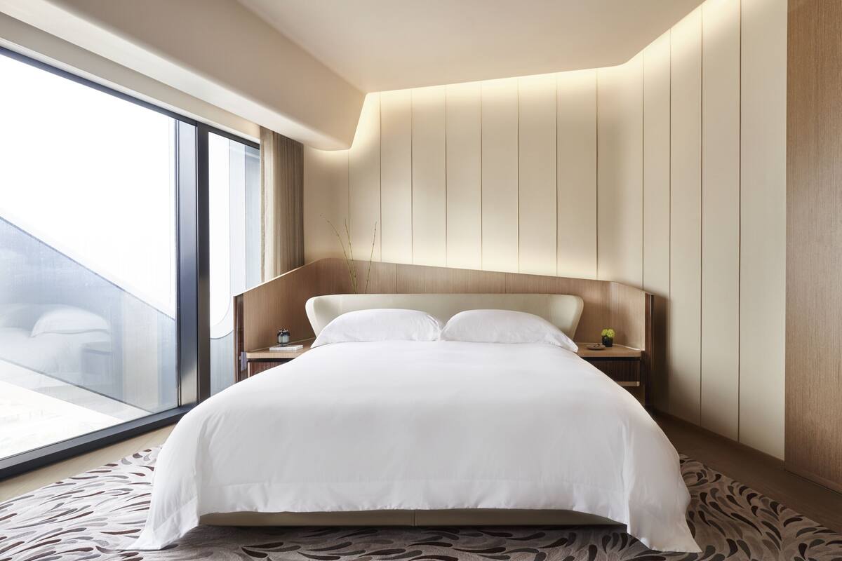 A modern hotel room with a large bed, a nightstand, a window with a view, and a curved wall design.