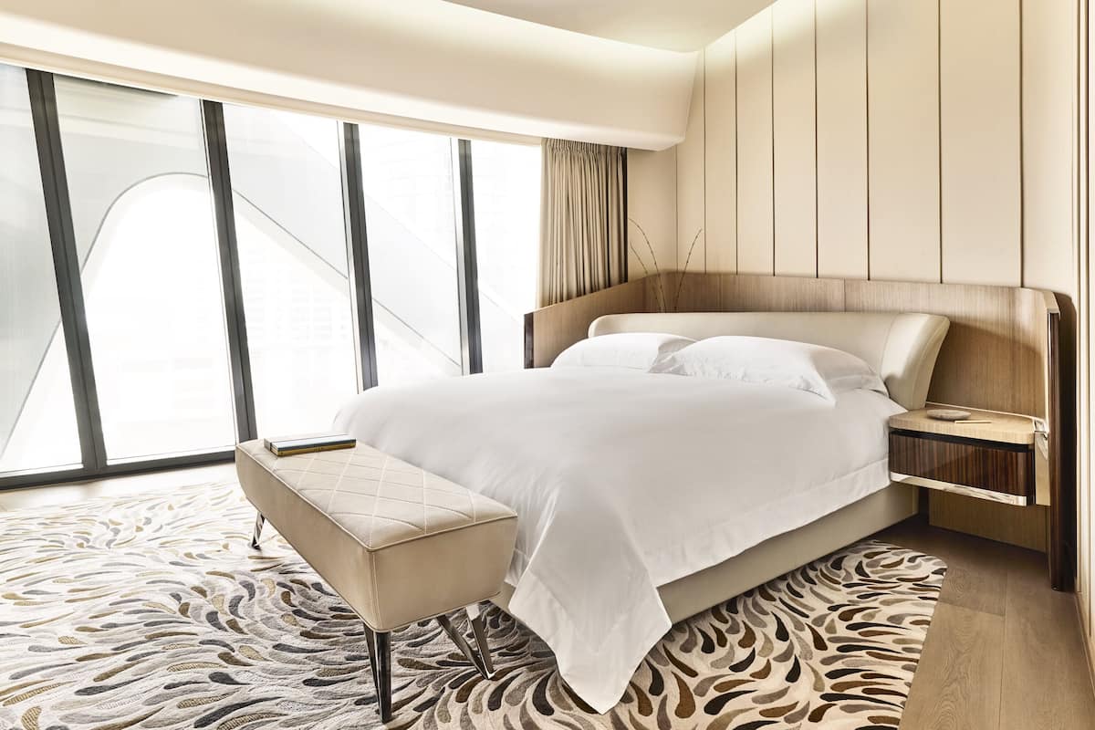 A modern hotel room with a large bed, a bedside table, a chair, and a view through floor-to-ceiling windows.