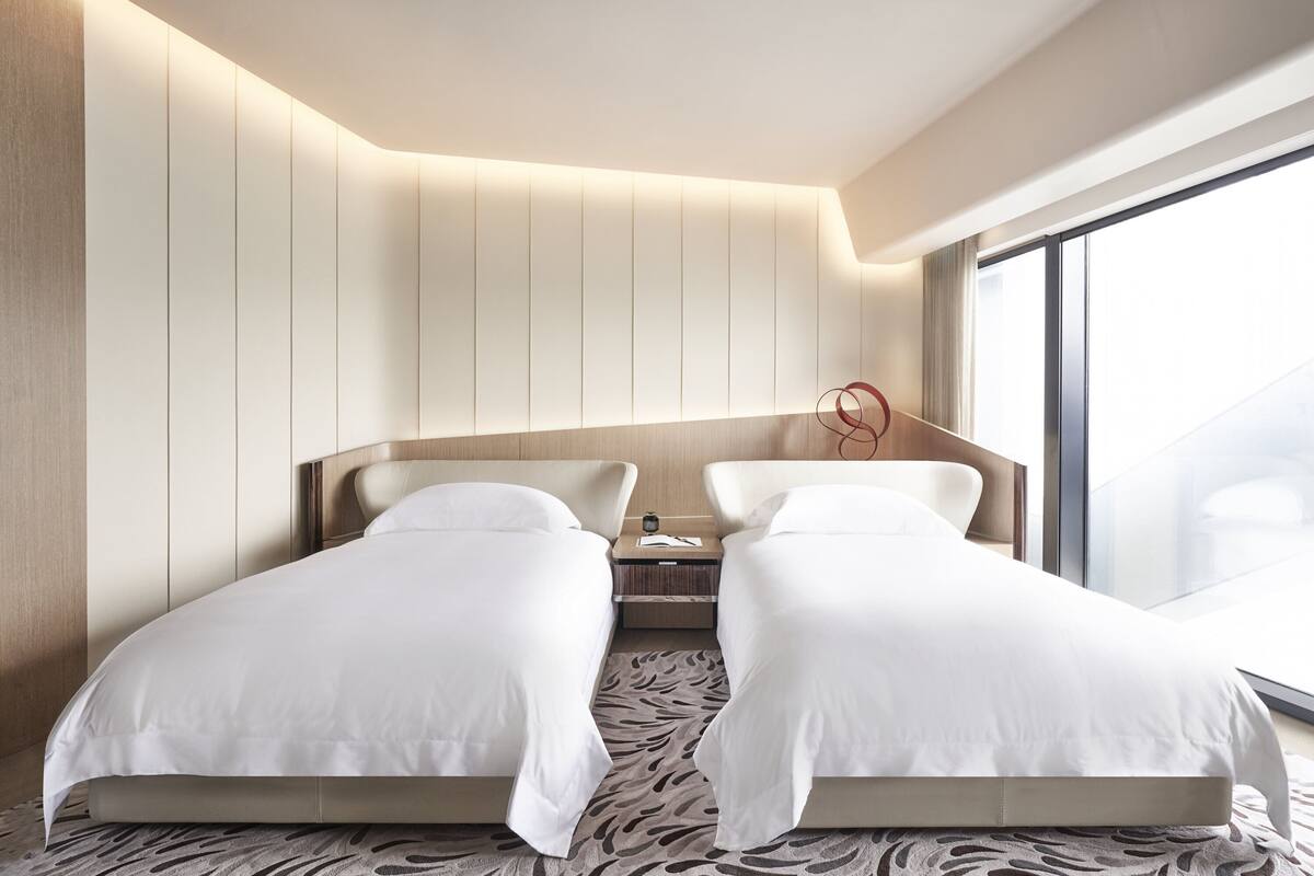 A modern hotel room with two single beds, a large window, and a unique ceiling design.