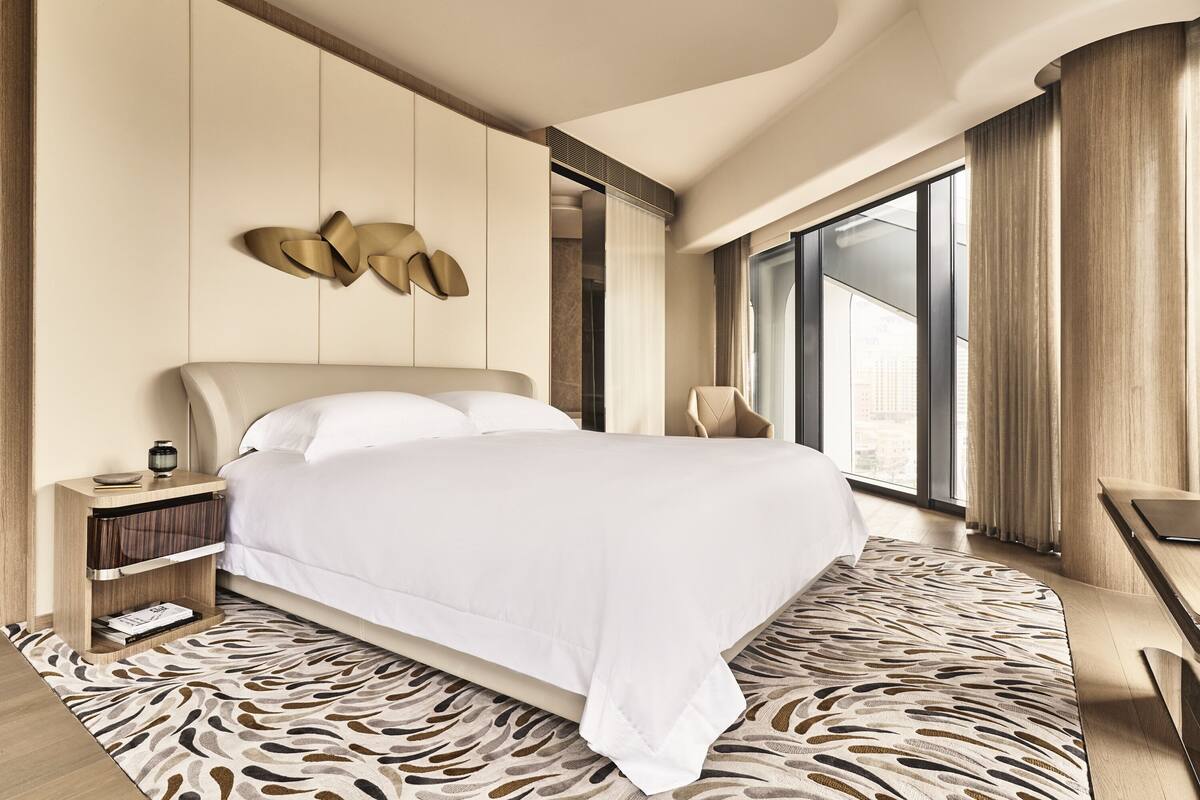 A modern hotel room with a large bed, a nightstand, a desk, and a view of the city through large windows.