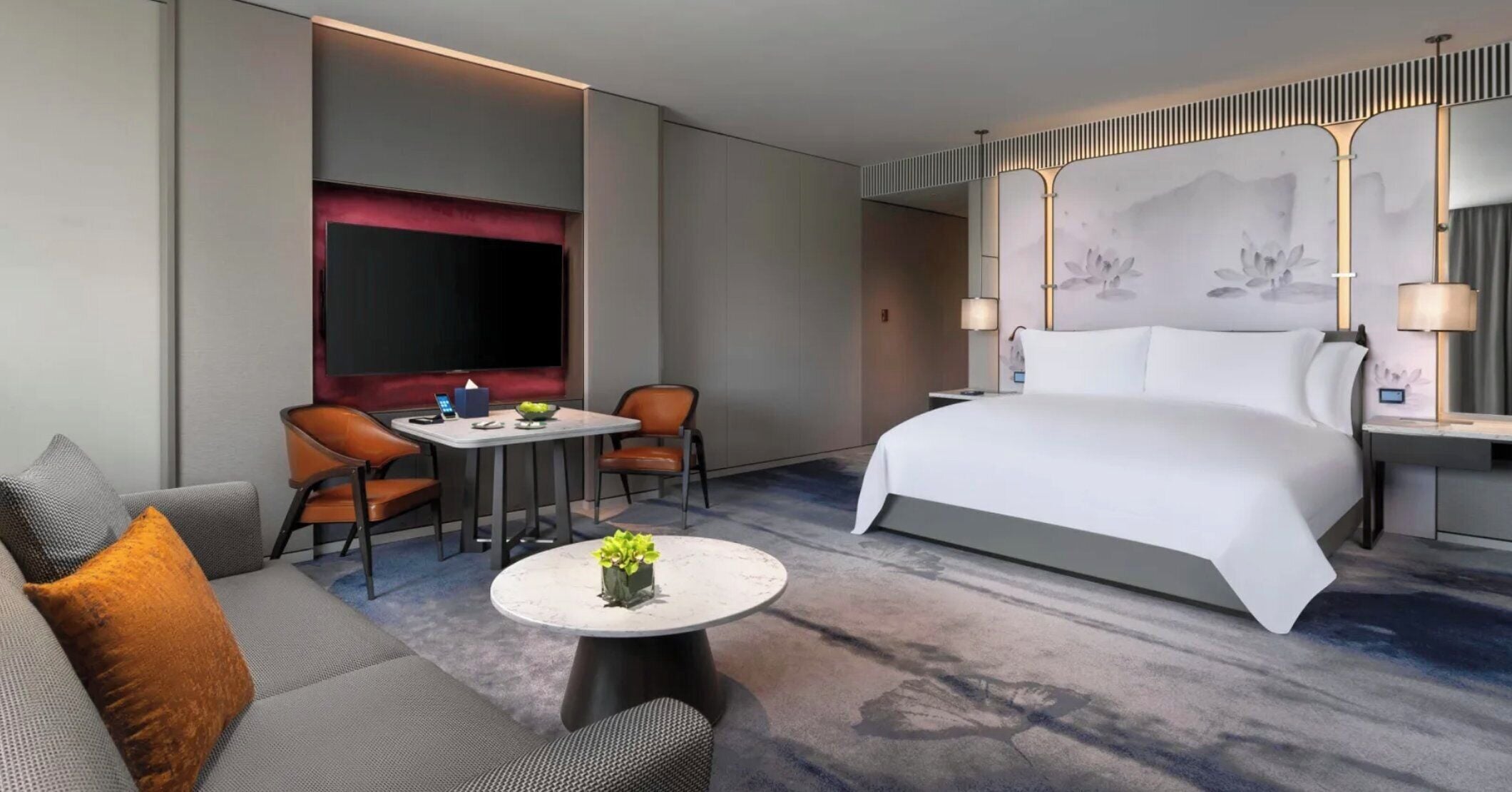 A modern hotel room with a large bed, a sofa, a small table with a plant, a desk, and a TV.