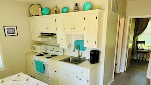 A compact kitchen with white cabinets, a stove, a microwave, and a coffee maker. There is a window with curtains and a chair in the adjacent room.