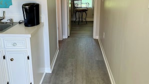 A hallway with a kitchen area on the left, a dining area with a chair and table, and a window with curtains.