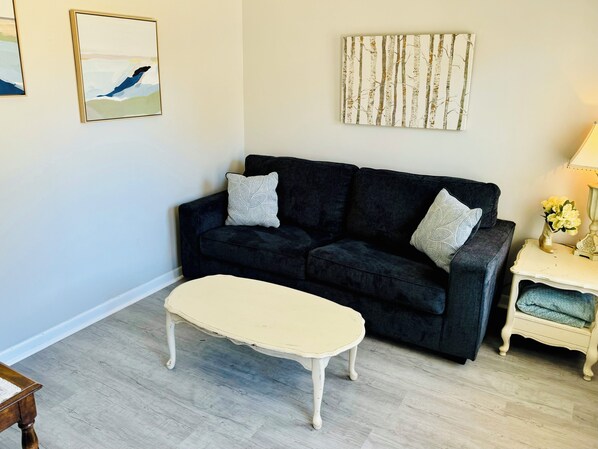 A living room with a dark sofa, a cream-colored ottoman, a small side table with a lamp, and a wall-mounted painting.