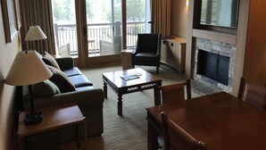 Habitación de hotel con sofá, sillón, mesa de comedor, televisor, chimenea y balcón con cortinas.