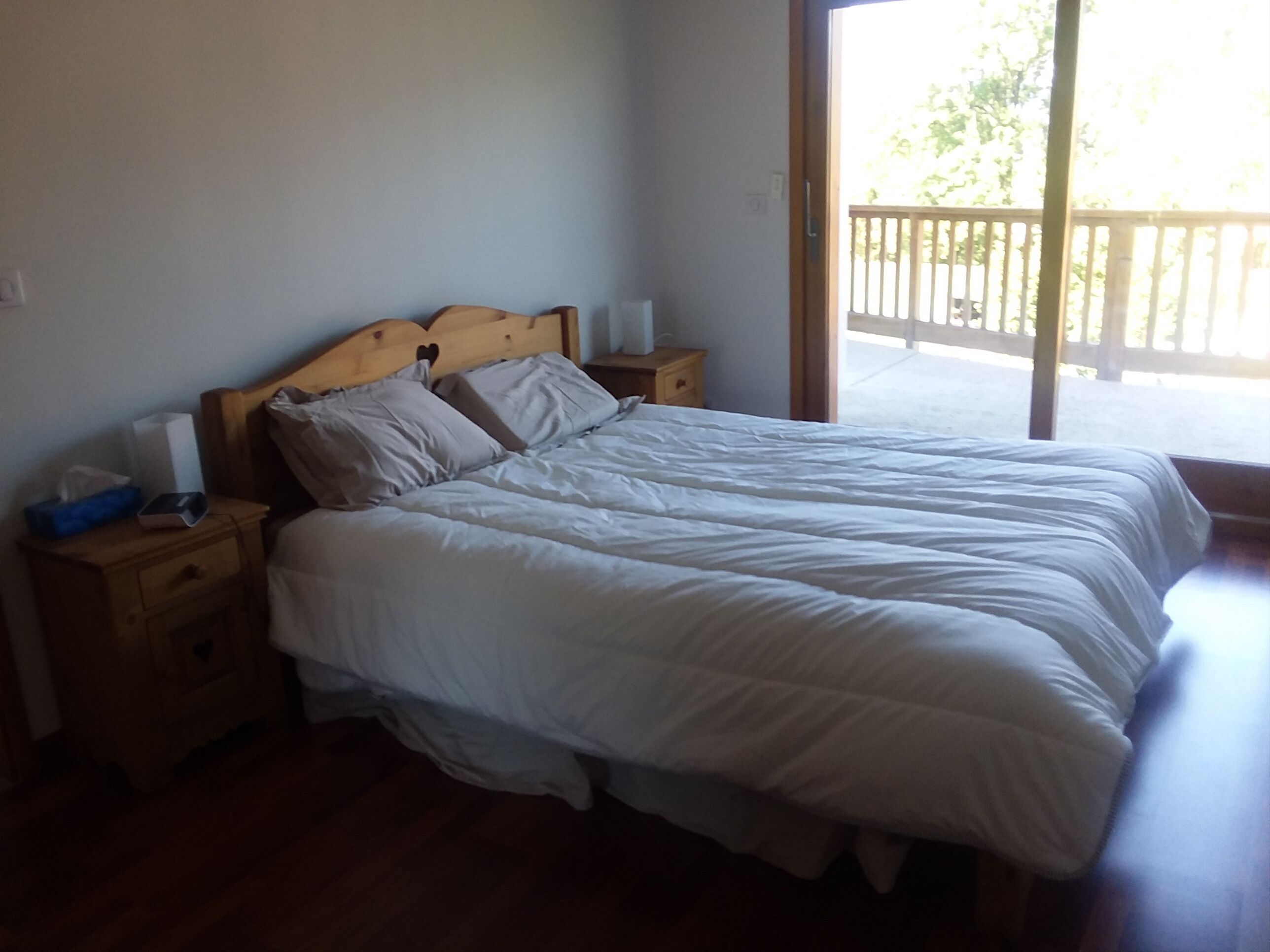 A bedroom with a wooden bed frame, white bedding, a nightstand with a tissue box, and a view of a balcony.