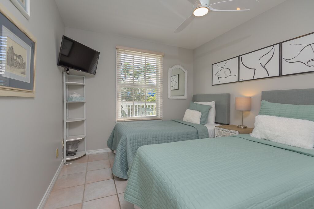 A bedroom with two beds, a TV, a window with blinds, a ceiling fan, and artwork on the wall.
