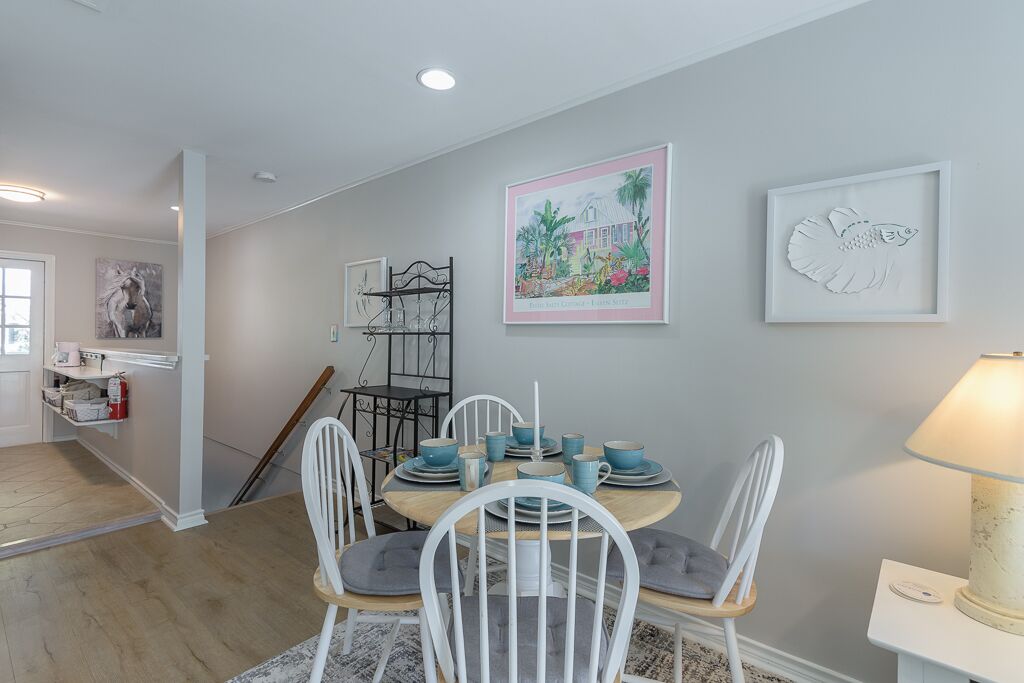 A dining area with a round table set for four, surrounded by white chairs with grey cushions. There are framed artworks on the walls, a white lamp on a side table, and a door leading to another room.