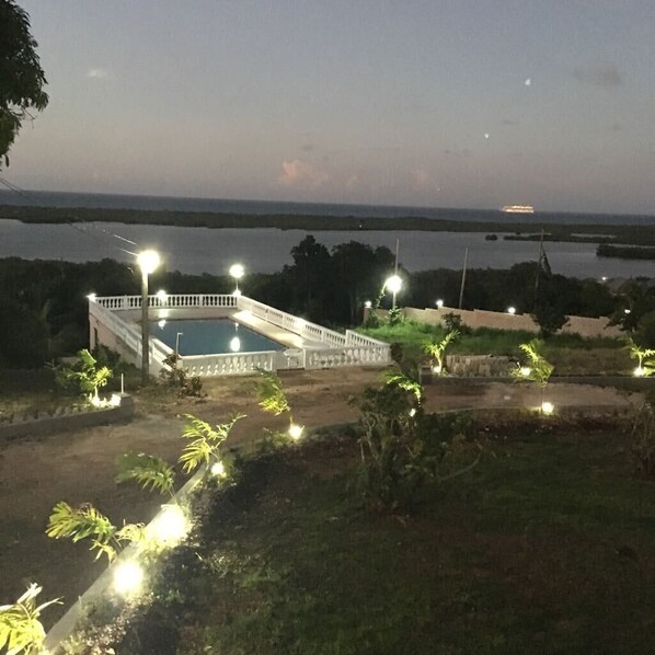 Una piscina con muelle blanco y un puente, rodeada de vegetación y farolas.