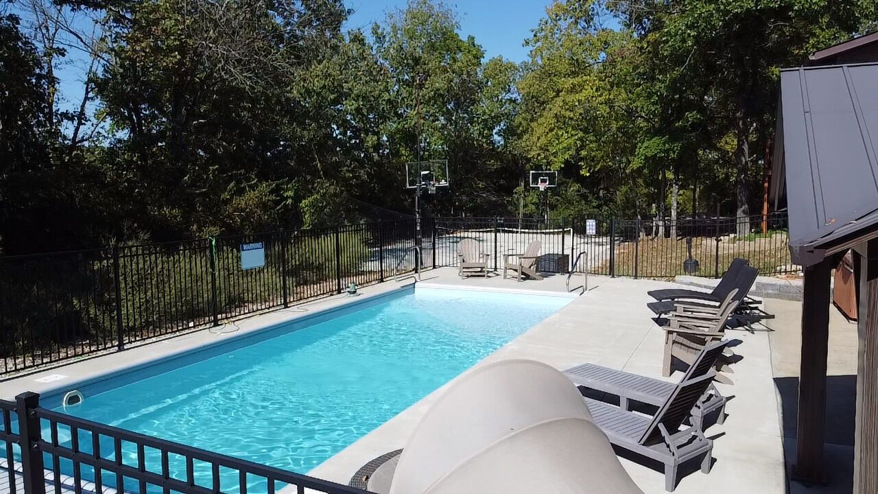 A swimming pool with a black fence, lounge chairs, and a basketball hoop.