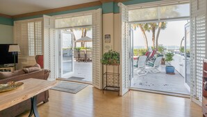 Un vaste salon comprenant une table basse en bois, un canapé et un téléviseur. De grandes fenêtres équipées de volets offrent une vue sur la terrasse extérieure, qui comprend une table à manger et des chaises.