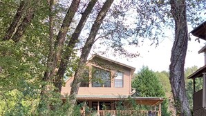 A wooden cabin with a porch overlooking a river, surrounded by dense foliage.