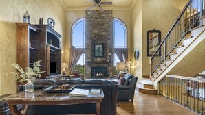 A spacious living room with a stone fireplace, leather sofas, a wooden coffee table, and a staircase with wrought iron railings.