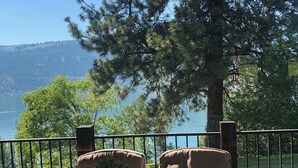 Un patio con dos sillones acolchados y una mesa de vidrio, con vista a un lago y una colina boscosa.