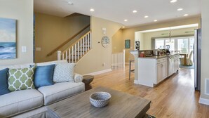 A spacious living room with a wooden coffee table, a beige sofa with patterned cushions, a staircase, and a kitchen area with white cabinets and a wooden countertop. - Brand New! 4-Br Bayside Townhome, Water and Pool View, Beach 1 block,  Free WiFi (Ocean City)