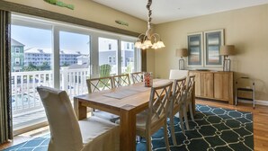 A dining area with a wooden table and chairs, a chandelier, and a view of buildings through large windows. - Brand New! 4-Br Bayside Townhome, Water and Pool View, Beach 1 block,  Free WiFi (Ocean City)