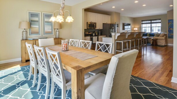 A spacious dining area with a wooden table and white chairs, a kitchen in the background, and a view of the living room. - Brand New! 4-Br Bayside Townhome, Water and Pool View, Beach 1 block,  Free WiFi (Ocean City)