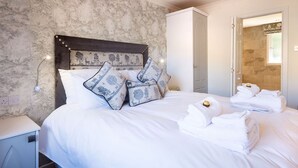 A neatly made bed with white linens and decorative pillows, a bedside table with a lamp, and a bathroom visible through an open door. - Deerstalker. Beautiful 5 star, 1 bedroom chalet sleeps up to 2 (Crianlarich)