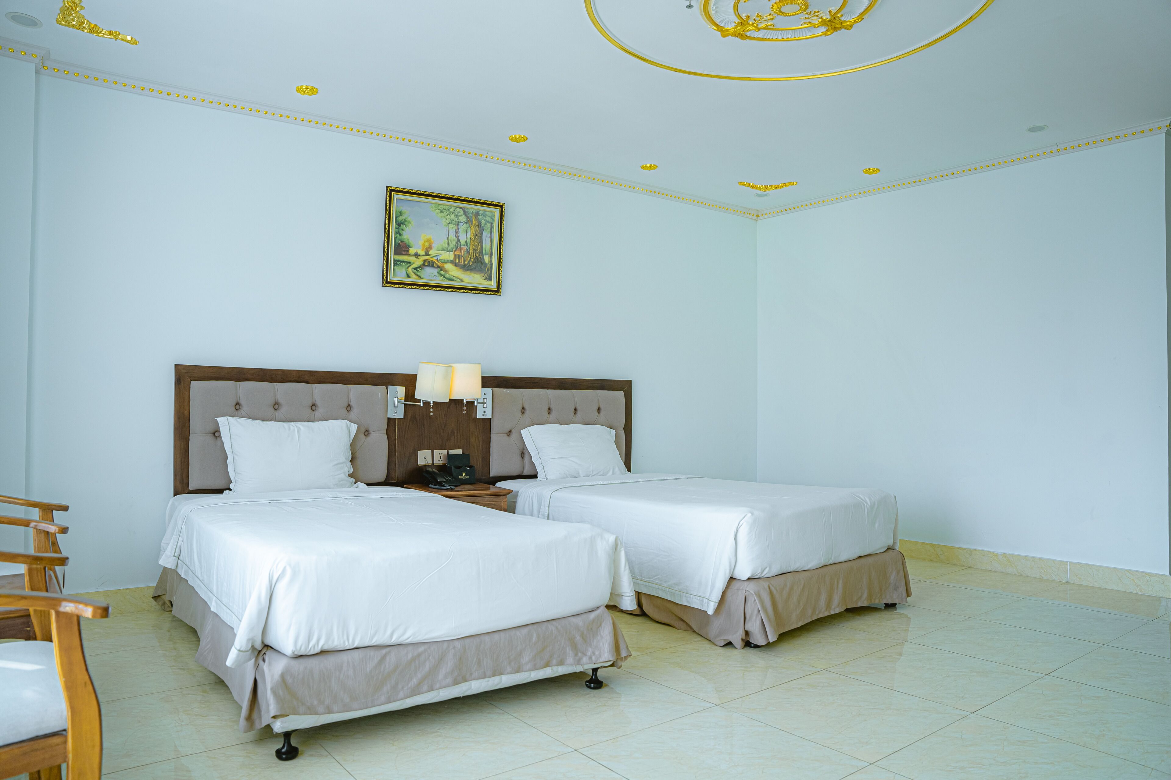 A hotel room with two beds, a wooden headboard, a nightstand with a lamp, a painting on the wall, and a tiled floor.