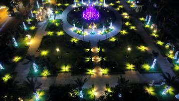 An aerial view of a well-lit garden with a central fountain, surrounded by pathways and illuminated plants.