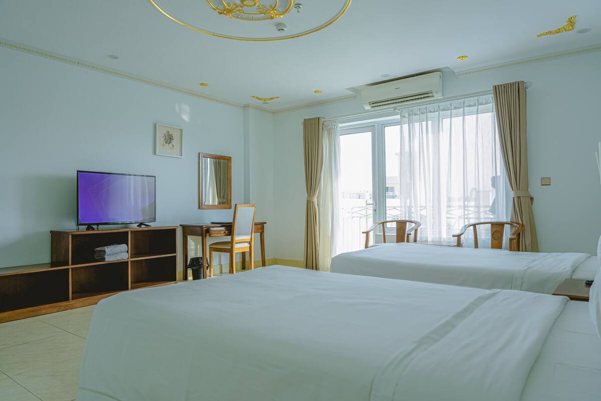 Ein Hotelzimmer mit zwei Betten, einem Schreibtisch mit Stuhl, einem Fernseher und einem großen Fenster mit Vorhängen.
