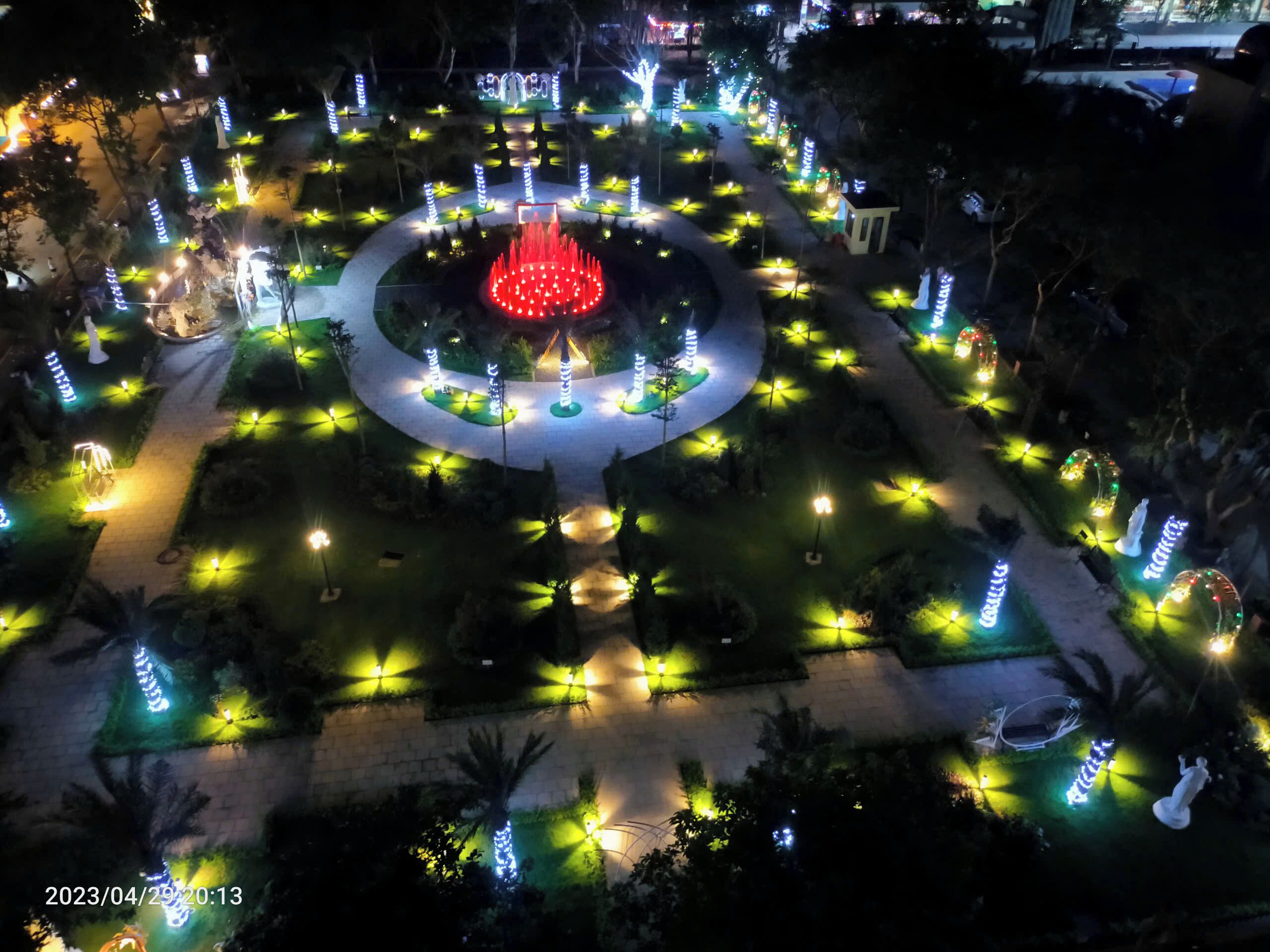 An aerial view of a well-lit park with a central fountain, surrounded by evenly spaced trees and illuminated pathways.