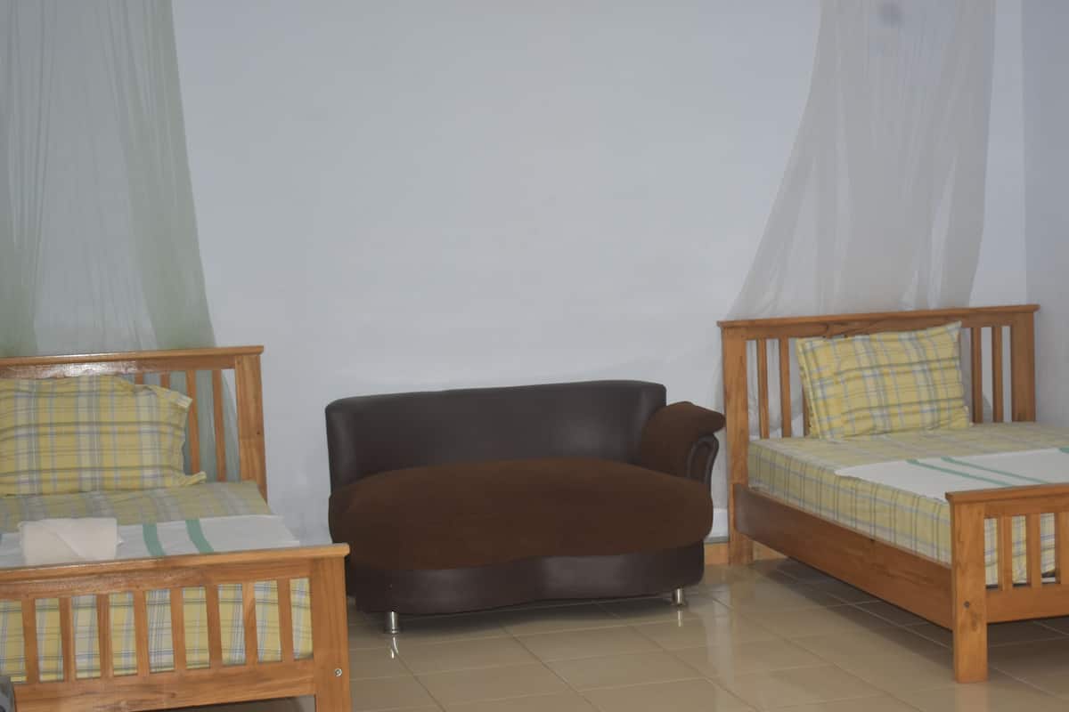 A room with two wooden beds, one with a yellow and blue checkered bedspread and the other with a yellow and white checkered bedspread. There is a brown sofa in the center of the room.