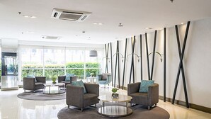 A modern lobby with grey sofas, a round coffee table, and a large window offering a view of greenery. - Jericho's Place at Sea Residences (Pasay)