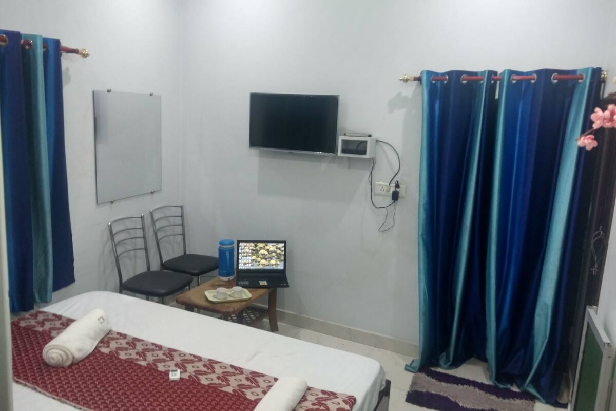 A hotel room with a bed, a desk with a laptop, a chair, a TV mounted on the wall, and curtains.