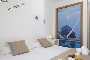 A hotel room with a bed, pillows, a bedside table with a lamp, a tray with breakfast items, and a large window with a view of a building and a body of water. - Singular Stays Oceanográfico (Valencia ciudad)