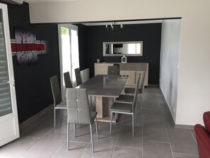 Un espace repas moderne avec une longue table et des chaises, un coin cuisine doté d’un comptoir et de placards, et un grand miroir accroché au mur.