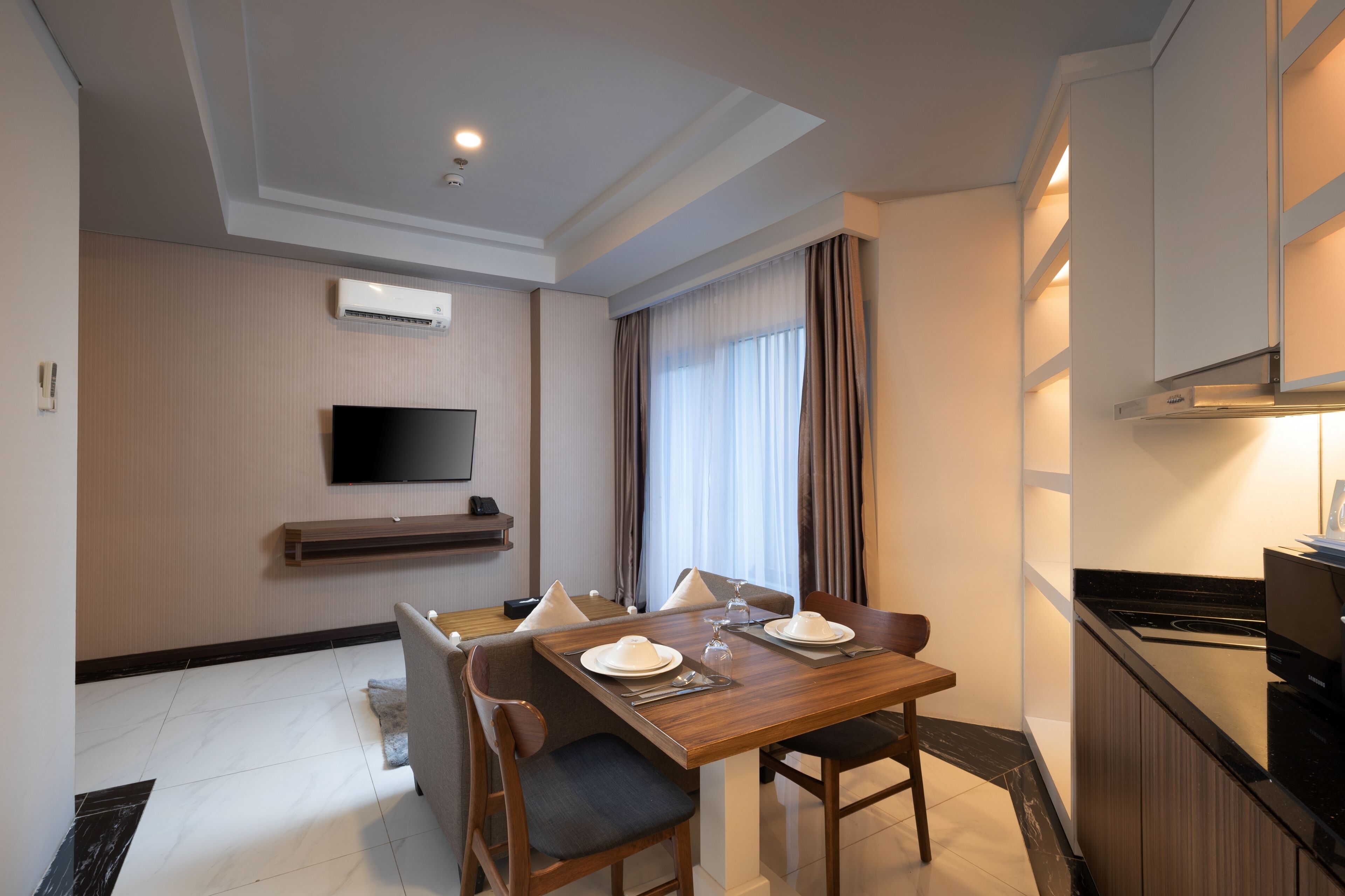 A hotel room with a dining area, a flat-screen TV mounted on the wall, a kitchen area with a stove, and a window with curtains.