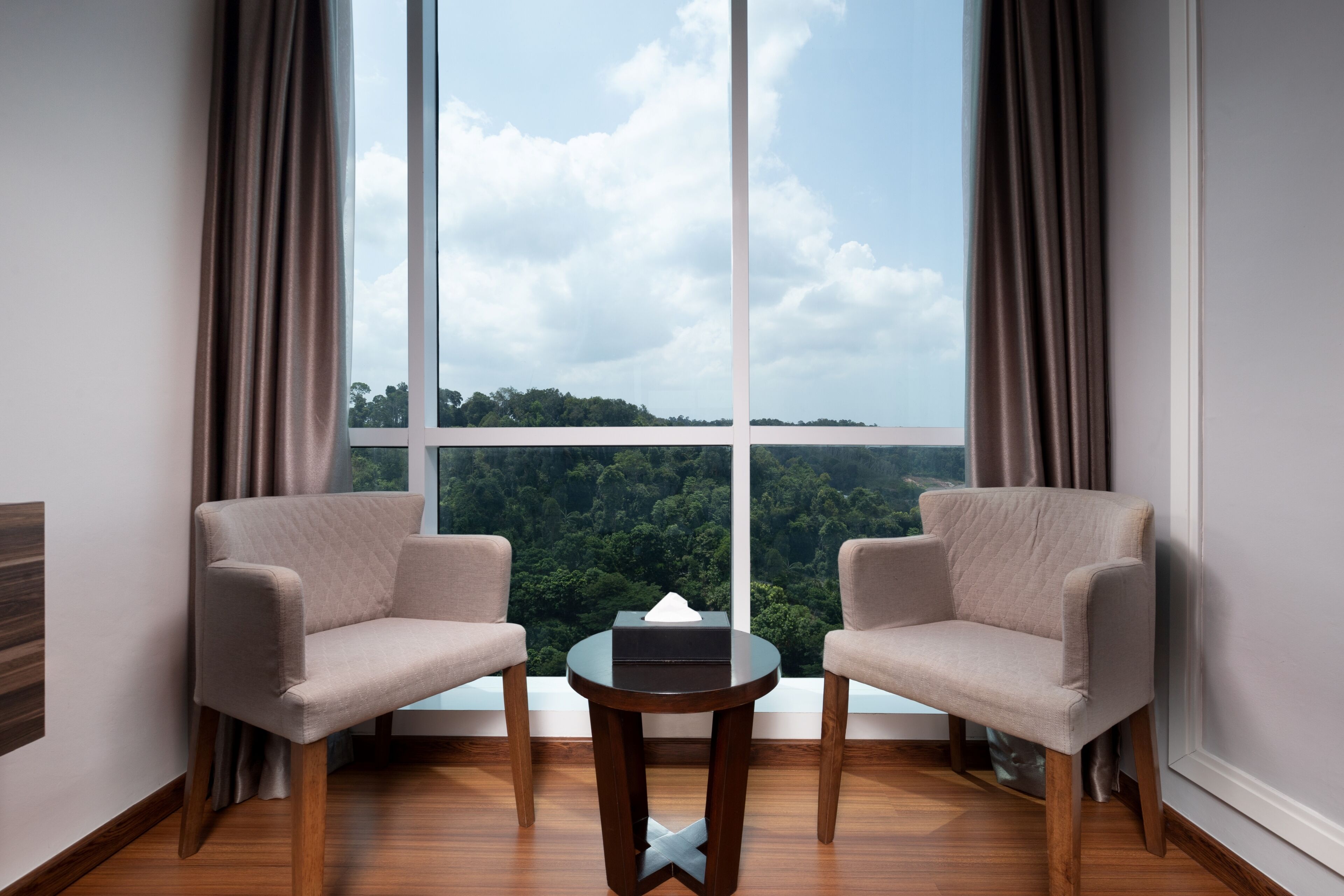 A room with two armchairs, a small round table, and a large window offering a view of a forested area.
