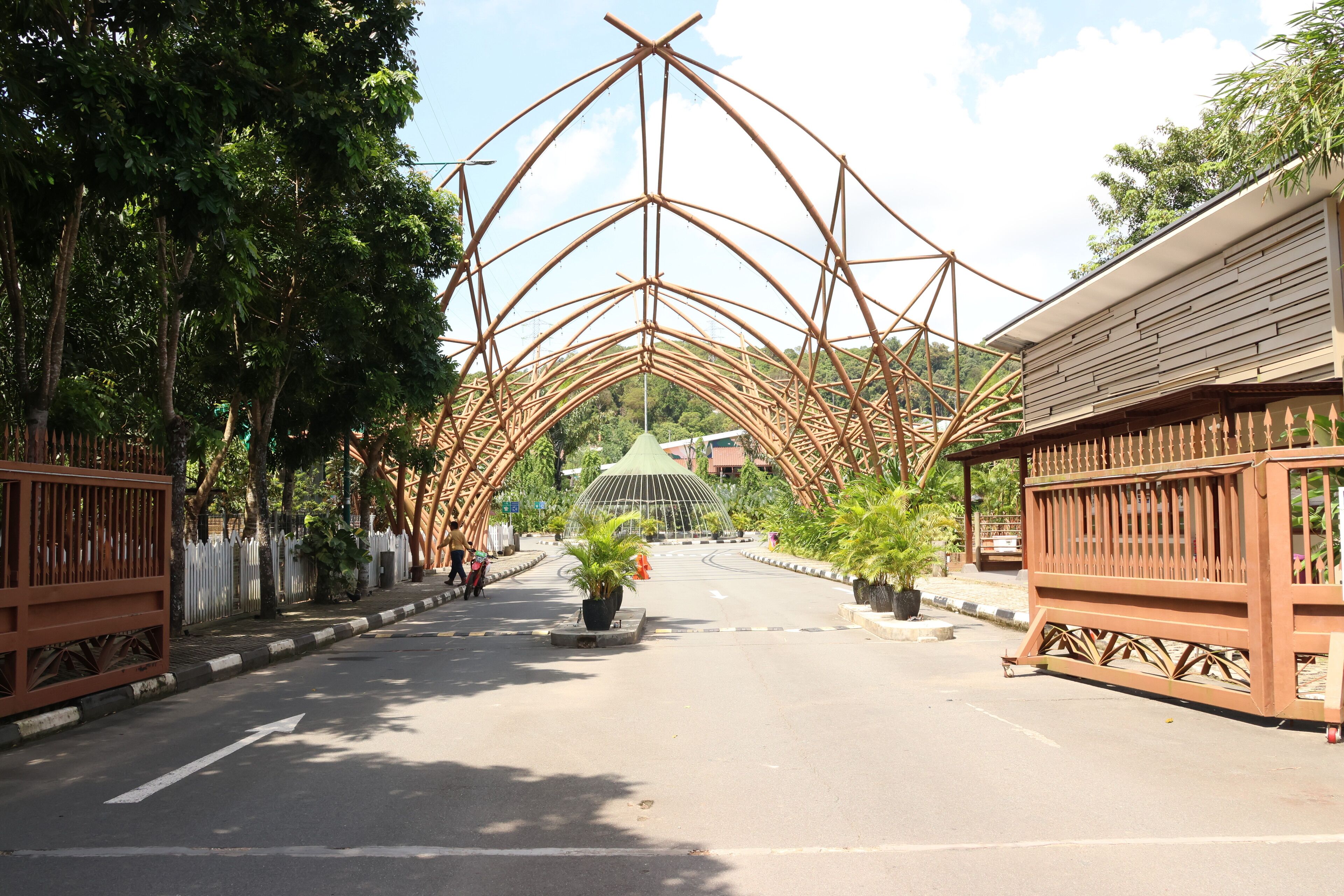 A street with a bamboo archway leading to a building with a green roof.