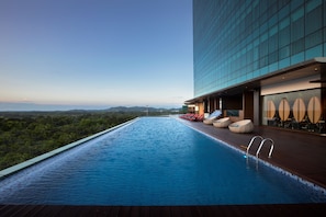 A rooftop infinity pool with a view of the mountains, lounge chairs, and a glass building.