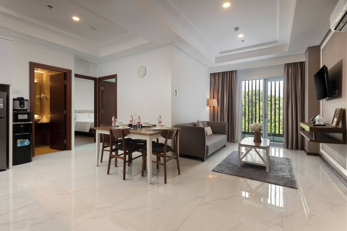 Una habitación de hotel moderna con comedor, sofá, televisión y balcón con cortinas.