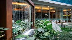 Un edificio moderno con fachada de vidrio, un elemento decorativo de agua en cascada y abundante vegetación.