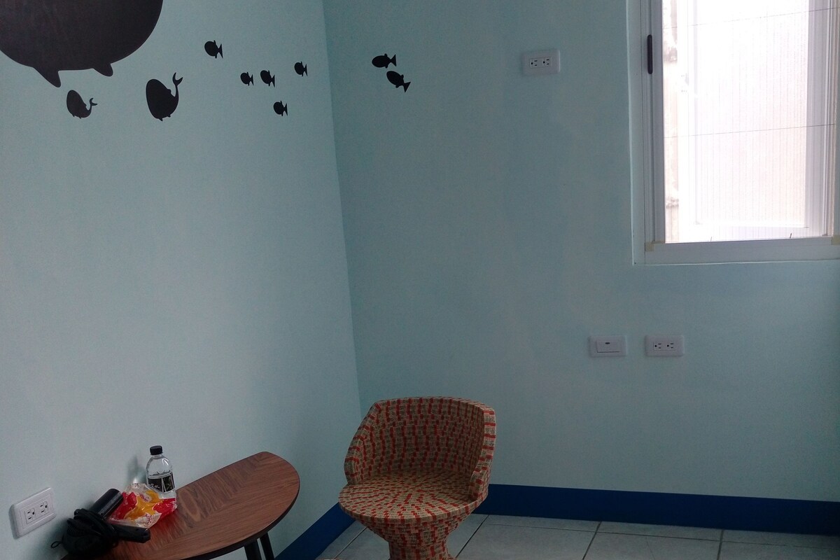 A room with a bed, a small table, a patterned chair, and a wall decal of a whale and fish.