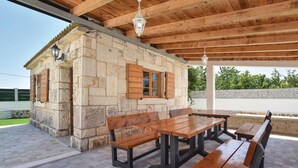 Een gebouw van steen met houten luiken, een betegeld terras en een houten tafel met bankjes.