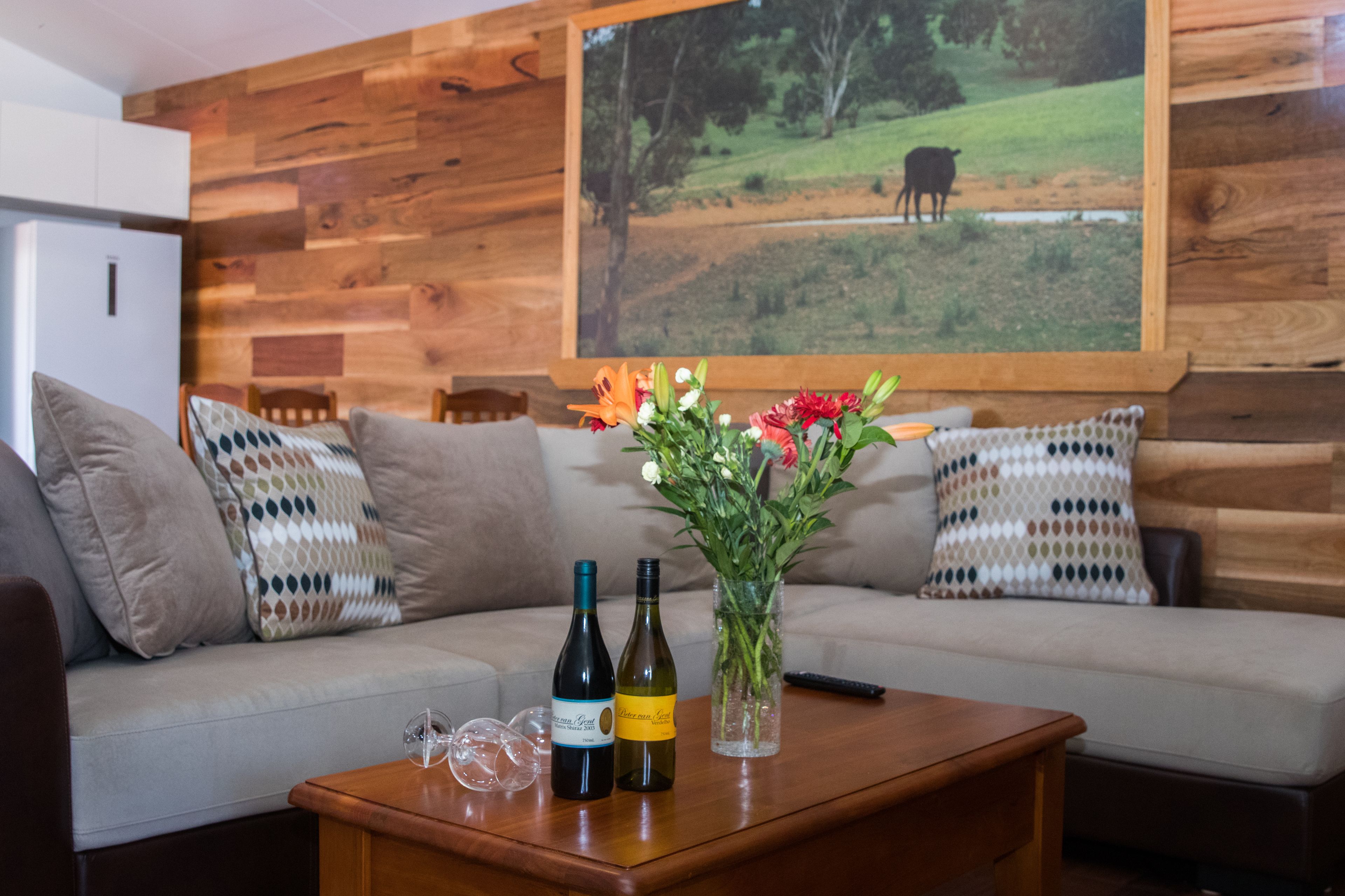 Un salón con pared de madera, un sofá con cojines estampados, una mesa de centro con botellas de vino y un florero con flores.