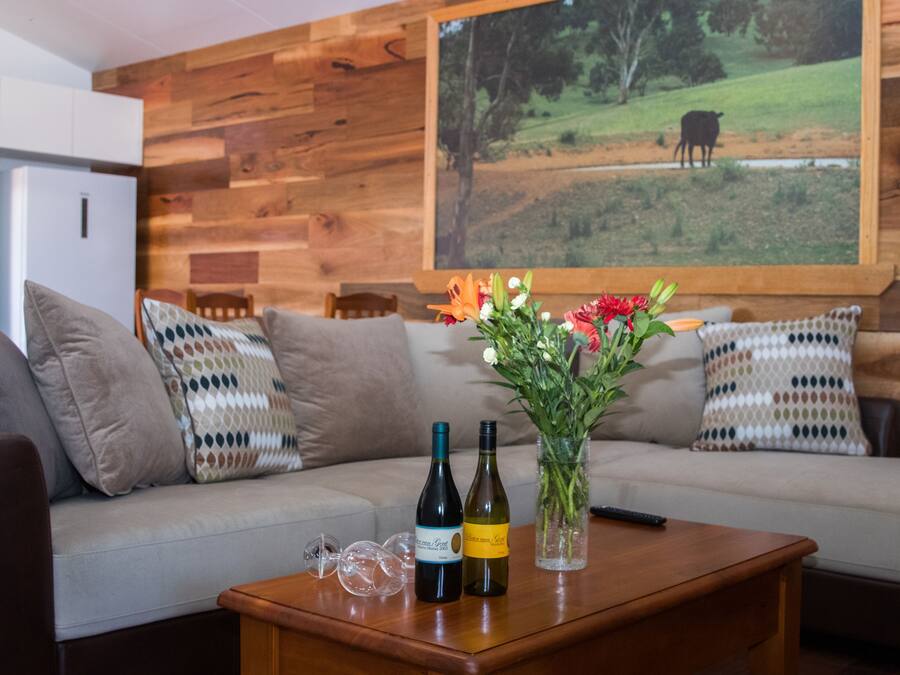 Un salón con pared de madera, un sofá con cojines estampados, una mesa de centro con botellas de vino y un florero con flores.