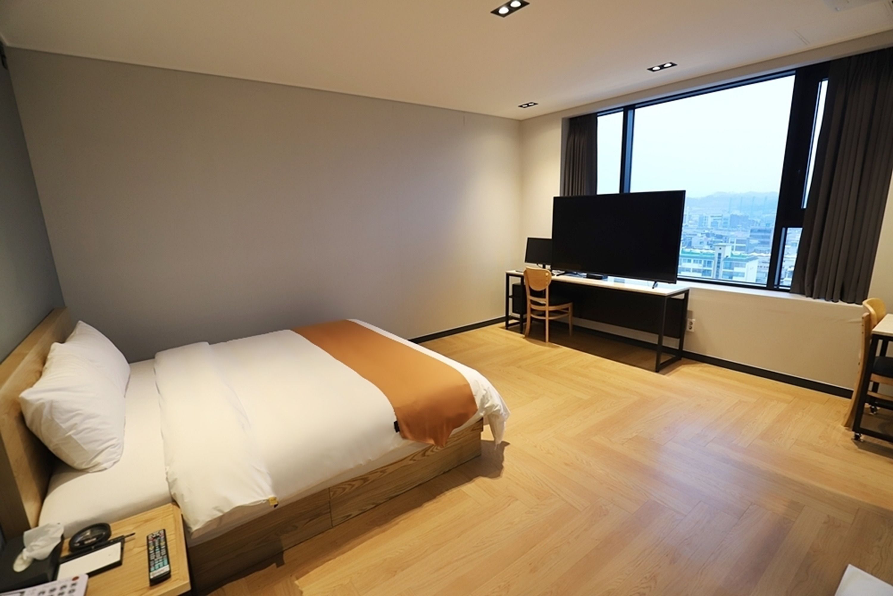 A hotel room with a bed, a desk with a TV, a chair, and a view of the city.