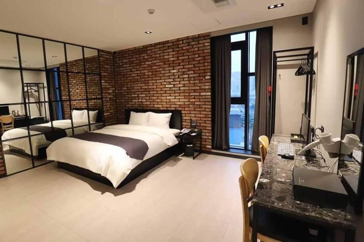 Une chambre d’hôtel moderne avec un mur en briques, un grand lit, un bureau avec un ordinateur et une vue par la fenêtre.