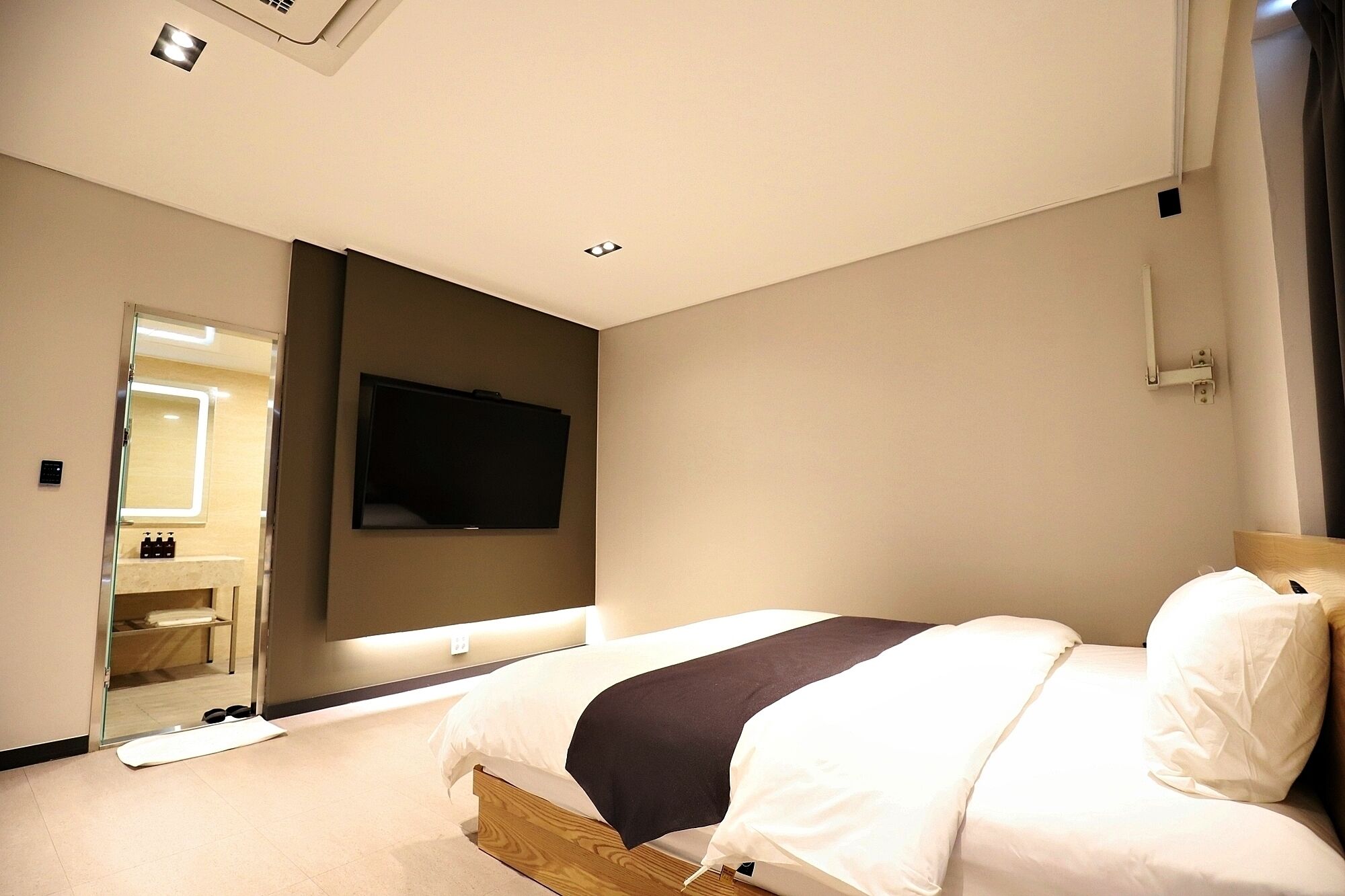 Habitación de hotel moderna con cama, televisor de pantalla plana en la pared, espejo y un estante pequeño con artículos de tocador.