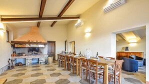 Un vaste espace de salle à manger avec une longue table et des chaises en bois, une cheminée en briques et une cuisine moderne équipée d’un climatiseur.