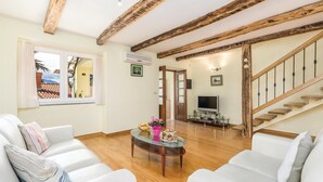 Una sala de estar con sofás blancos, una mesa de centro de cristal, vigas de madera en el techo, un televisor y una escalera.