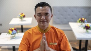A person in an orange uniform with a name tag, standing in a room with tables and floral centerpieces. - Front One Boutique Karawang (Karawang)