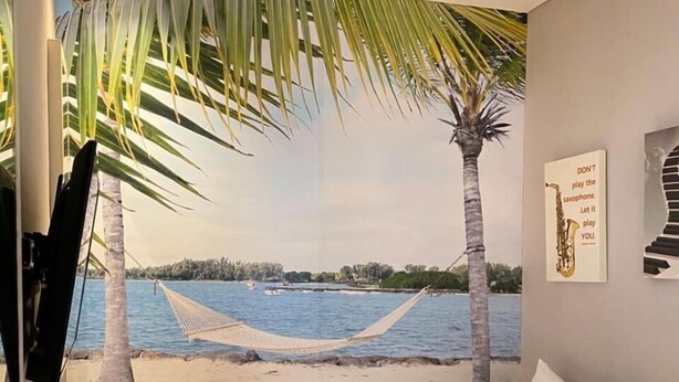 A hotel room with a bed, a TV, a desk with a laptop, and a view of a beach with palm trees and a hammock. - Front One Boutique Karawang (Karawang)