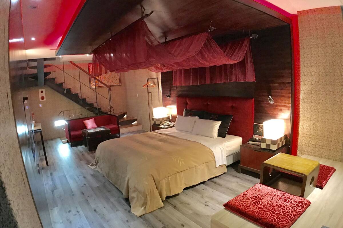 A modern hotel room with a large bed, a red sofa, a small table, and a staircase.
