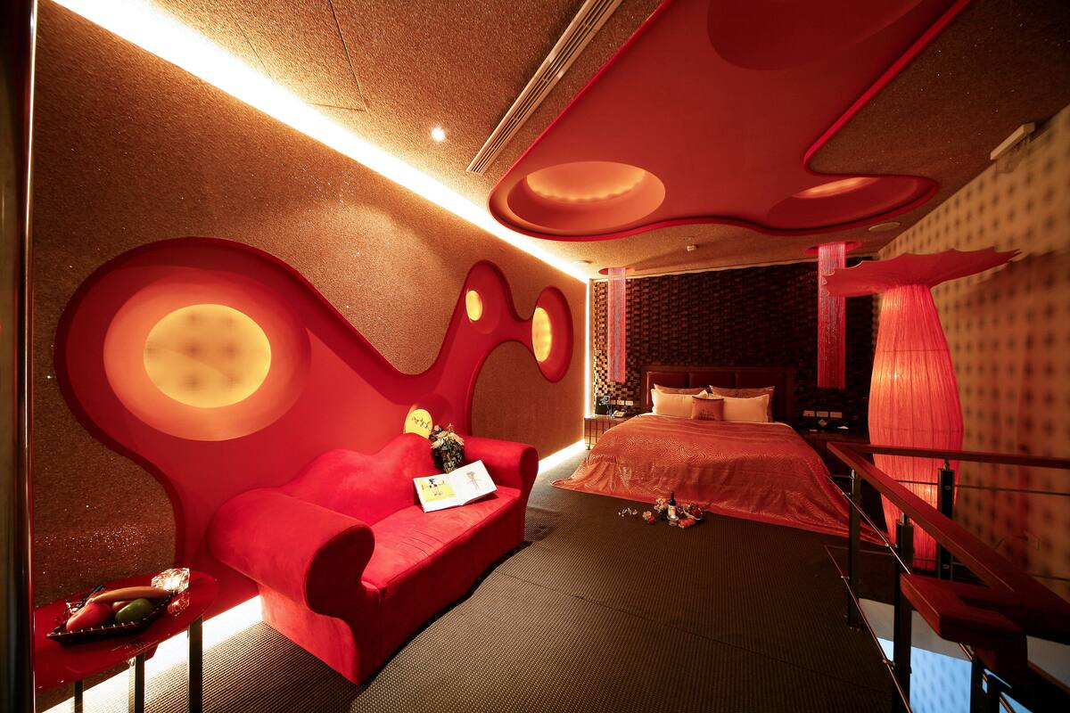 A modern hotel room with a red sofa, a bed with a red headboard, and a unique ceiling design.